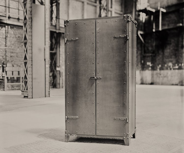 Schwarz-Weiss Aufnahme im Sepiaton eines soliden Stahlschranks mit Nieten rund um die Türen, der in einer industriellen Halle steht.