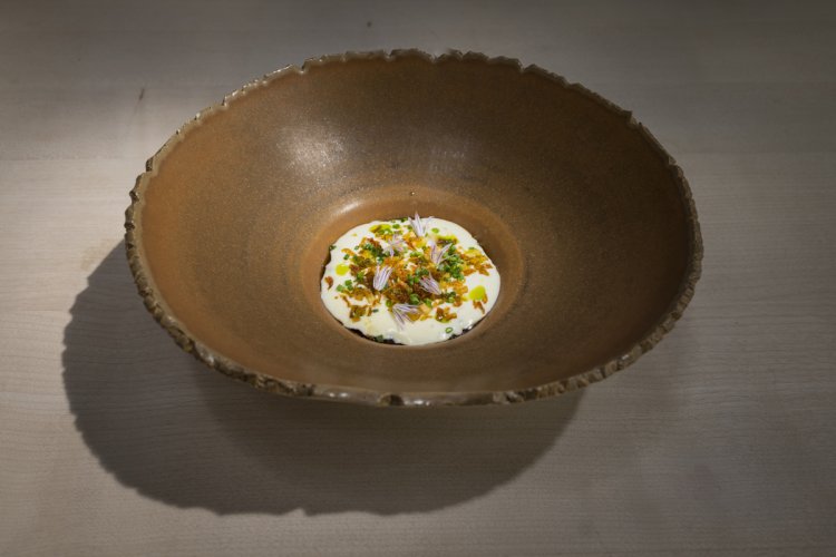 Brauner Teller mit weisser Speise in der Mitte, geschmückt mit gebratenen Zwiebeln, grünen Kräutern und violetten Blüten.