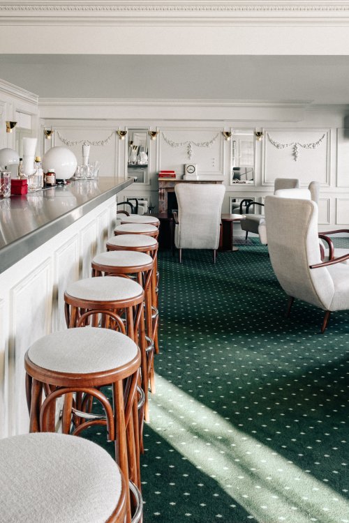 Hotelbar mit dunkelgrünem Teppich, weiss gepolsterten Barhockern und Sesseln und Stuckaturen an den weissen Wänden und Decken.