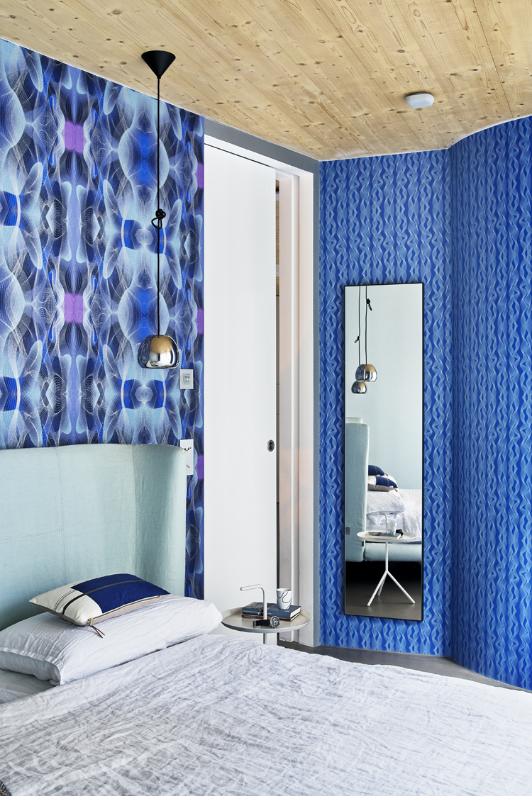 Schlafzimmer mit Bett im Vordergrund und blau gemusterten Tapeten im Hintergrund