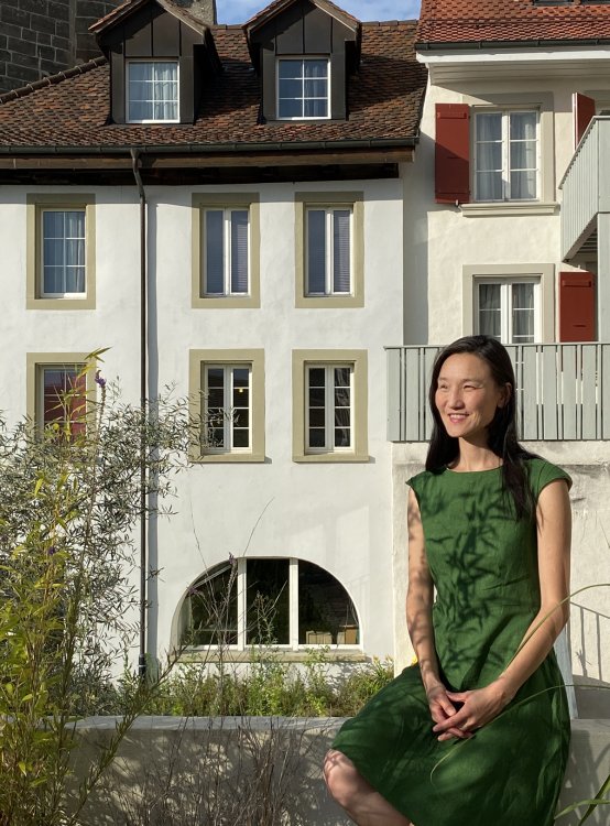 Saikal Zhunushova sitzt in einem grünen Kleid auf einer tiefen Gartenmauer, dahinter die Fassaden von Wohnhäusern, links im Bild ein grüner Busch