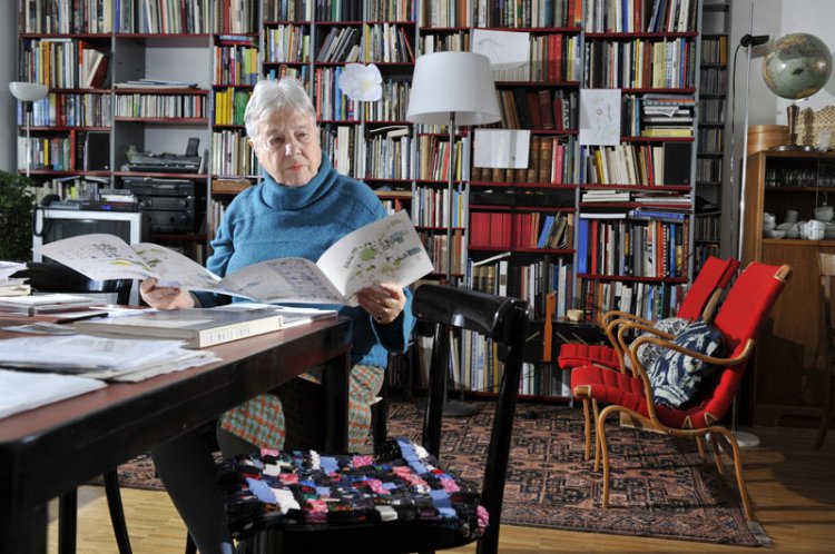 Verena Huber sitzt mit einer offenen Broschüre an ihrem Schreibtisch vor ihrem Bücherregal, rechts dahinter zwei rote bequeme Stühle