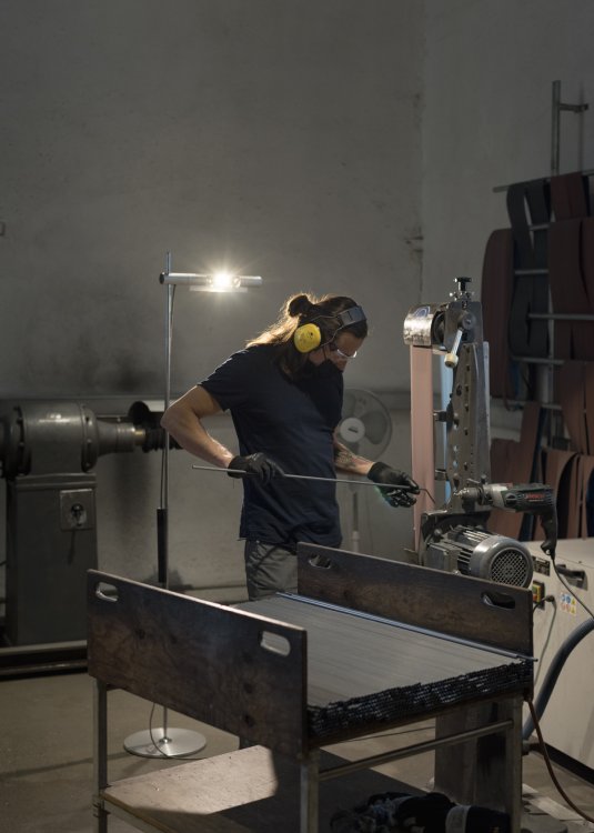Mann mit gelbem Gehörschutz und schwarzer Maske schleift ein Stahlrohr an einer Schleifmaschine, im Hintergrund eine Leuchte von Baltensweiler