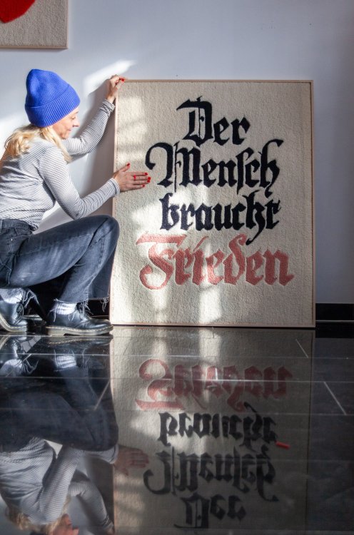 Frau mit blauer Mütze und blonden Haaren kniet seitlich neben einem Wandteppich mit dem Schriftzug Der Mensch braucht Frieden