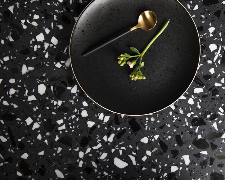 Eine schwarze Tischplatte in Terrazzo-Optik mit einem Teller mit goldenem Löffel und Pflanze.