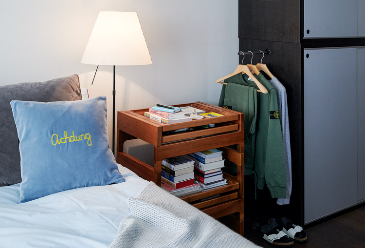 Detailansicht aus dem Schlafzimmer, Bettauschnitt mit hellblauem Kissen und gelber Stickerei, daneben Nachttisch aus Holz sowie ein paar Kleidungsstücke aufgehängt an einer Kleiderstange, die seitlich am Schrank befestigt ist.