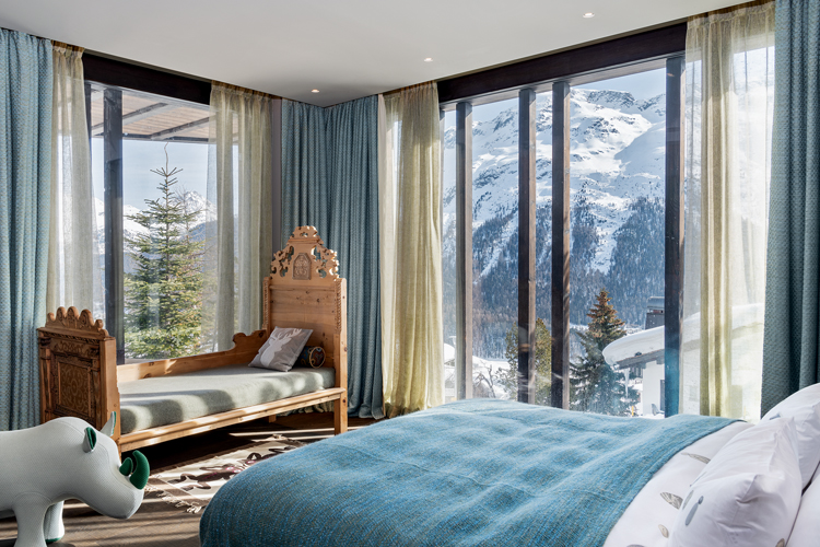 Im Schlafzimmer steht ein Bett mit hellblauer Tagesdecke, die Eckfenster gewähren einen Blick auf die Winterlandschaft.