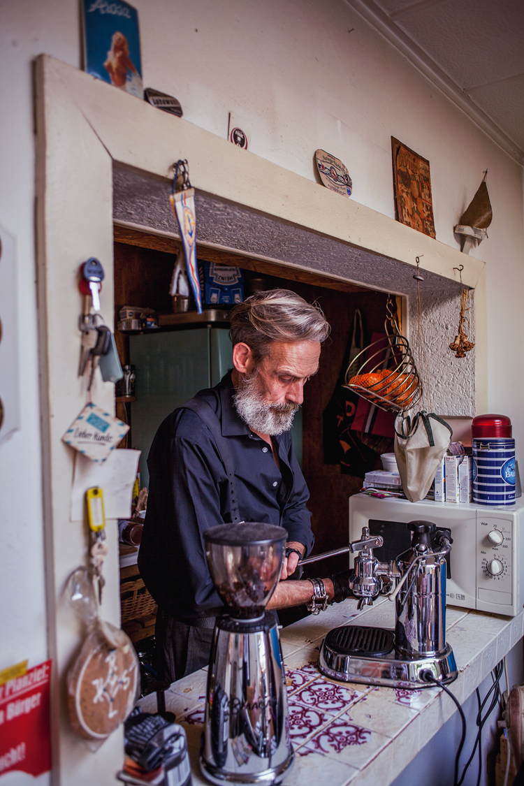 Mann mit grauem Bart steht hinter einer Küchentresen und macht einen Kaffee.