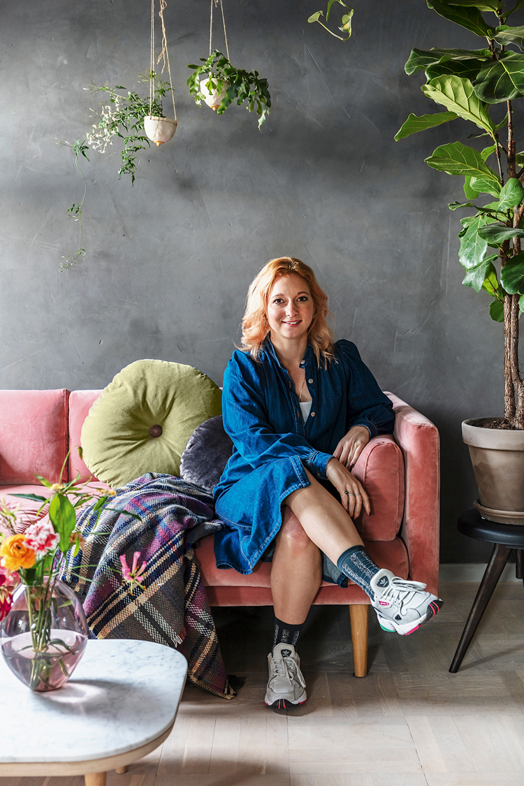Katharina Beckmann sitzt auf ihrem Sofa mit den Beinen überschlagen und lächelt in die Kamera.