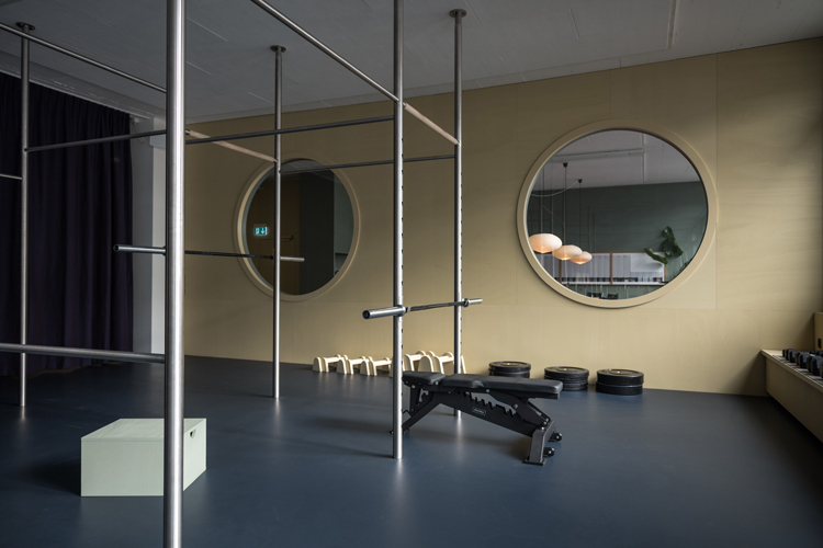 Sportraum mit diversen Geräten wie Reckstangen und Hantelbank, dunkelgrauem Boden und gelber Wand mit zwei parallel angelegten runden Fenstern in der Wand.