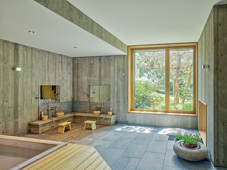 Schlichtes Badezimmer mit grauem Steinboden und Betonwänden und Raumhohem Fenster mit Blick in die grüne Natur.