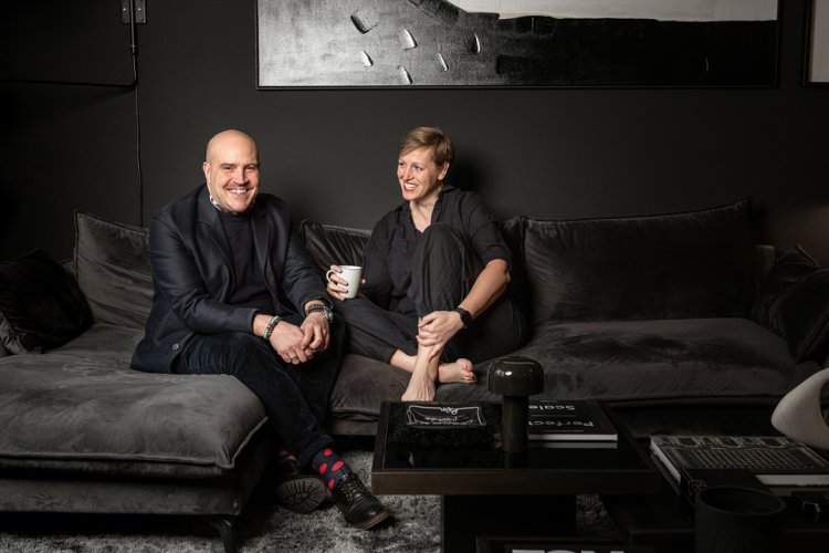 Das Paar Oliver W. Widmer und Lena Papasz sitzen nebeneinander lachend auf dem dunkelgrauen Samt-Sofa vor dunkelgrauer Wand.