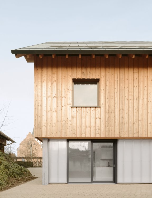 Abbgebildet ist die Seitenansicht des Hauses. Oben besitzt es eine vertikale Holzschalung und unten eine transparente Polycarbontfassade.