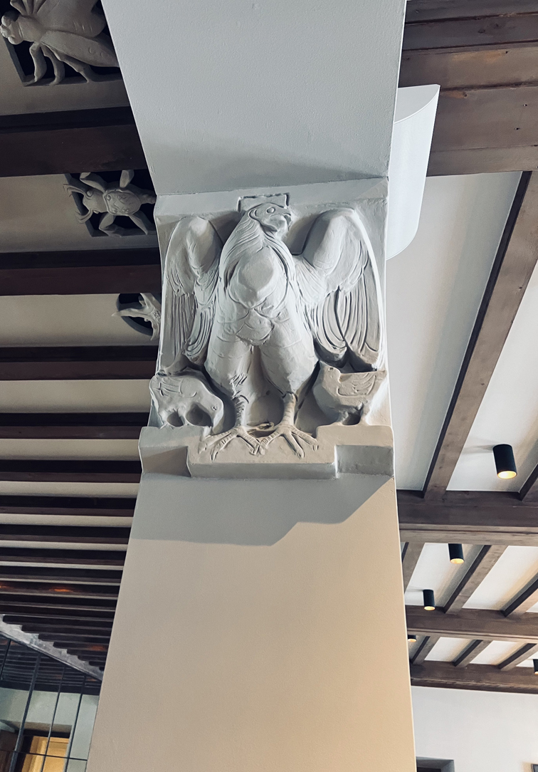 Detail einer Tierskulptur (ein Adler) von Wilhelm Schwerzmann als Abschluss einer Säule im Restaurant. Weitere Deckenverzierungen in Form von Tierskulpturen sind darüber sichtbar.
