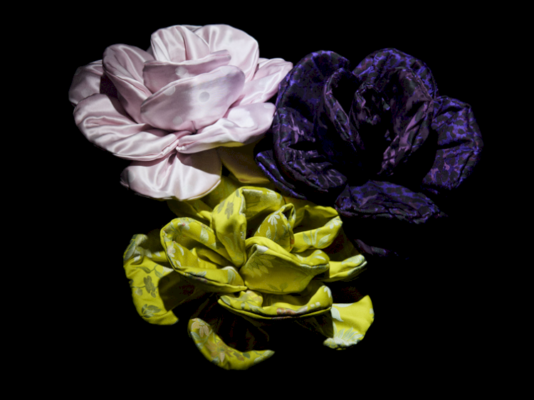 Drei Blüten aus Stoff gefertigt in verschiedenen Farben vor einem schwarzen Hintergrund, beleuchtet.