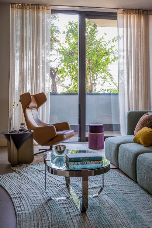 Ein Foto des Wohnzimmers. Im Wohnzimmer befindet sich ein brauneer Sessel, ein Beistelltisch aus Glas und ein blaues Sofa mit farbigen Kissen.