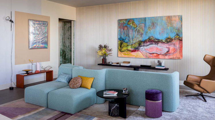 Ein Foto des Wohnzimmers von der anderen Seite. Zu sehen ist ebenfalls das blaue Sofa und brauner Sessel. Zu sehen ist noch ein schönes, farbiges Gemälde und ein paar Beistelltische.
