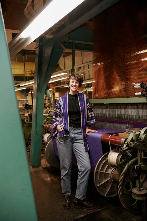 Portrait von Marie Schumann die vor einer Textilmaschine steht.