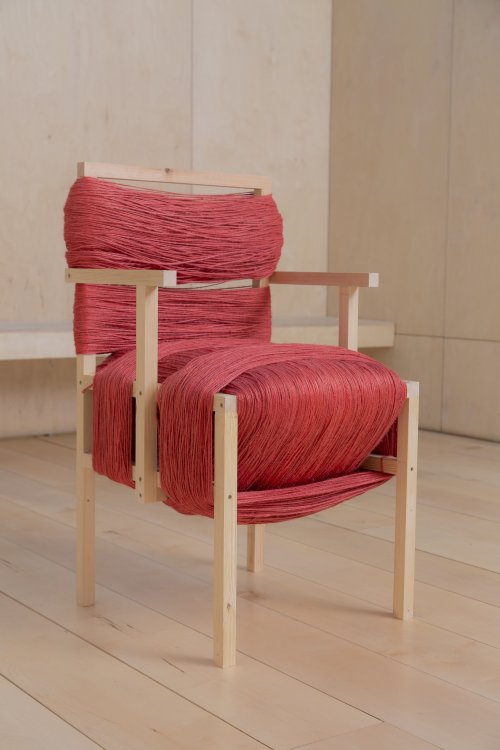 Ein einfacher Holzstuhl ist auf Sitz- und Lehnfläche etliche male mit dickem roten Garn umhüllt, was eine dicke Polsterung bildet.