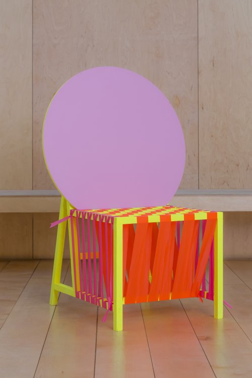 Ein knallig bunter Stuhl mit einer grossen, runden rosaroten Lehne und einem neongelb-gestrichenen Beingestell. Die Sitzfläche des Stuhls bilden leuchtend orange dicke Bänder, die sich um das Untergestell wickeln.