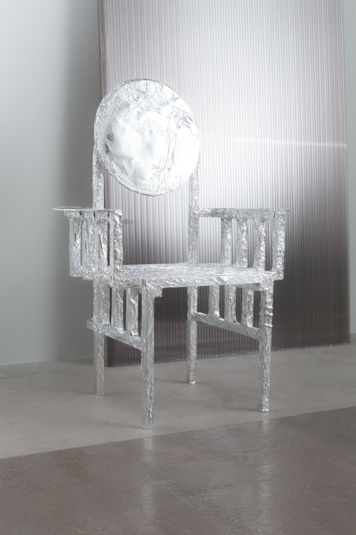 Ein Stuhl mit einer runden Rückenlehne ist vollkommen von silber-glänzender Alufolie umhüllt und reflektiert das auf ihn fallende Licht.