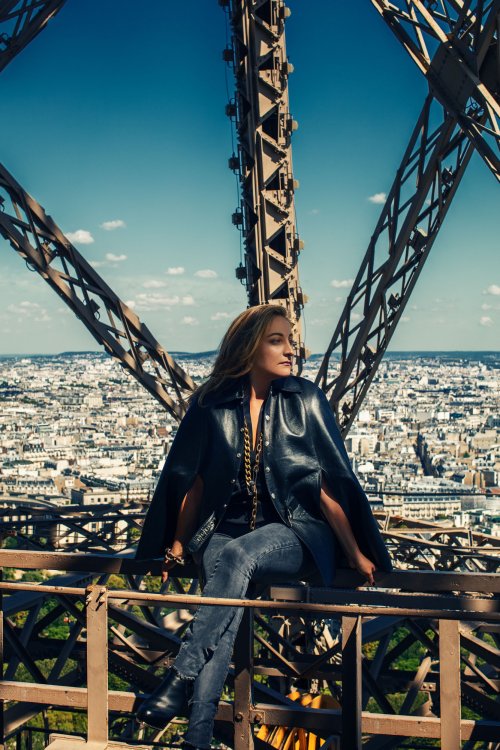 Die Architektin und Innenarchitektin Aline Asmar d'Amman in einer Lederjacke und Jeans auf dem Geländer des Eifelturms sitzend, dahinter die Aussicht über die Stadt Paris.