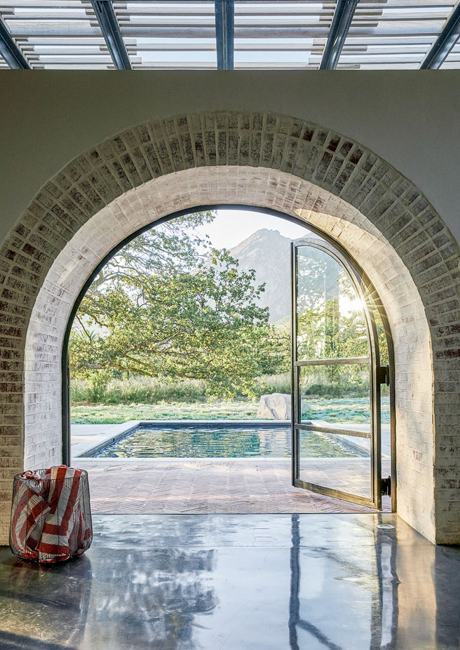 Ein runder Backsteinbogen gibt Sicht vom Innern des Hauses auf einen dahinterliegende Pool und den grünen Garten frei.