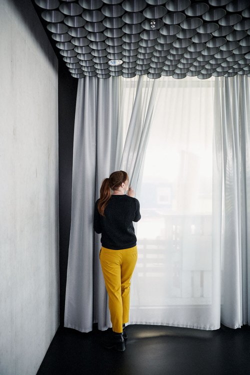 Die Textildesignerin Annette Douglas steht in schwarzem Pullover und senfgelber Hose an einem raumhohen Fenster mit einem hellgrauen transparenten Vorhang und blickt nach draussen.