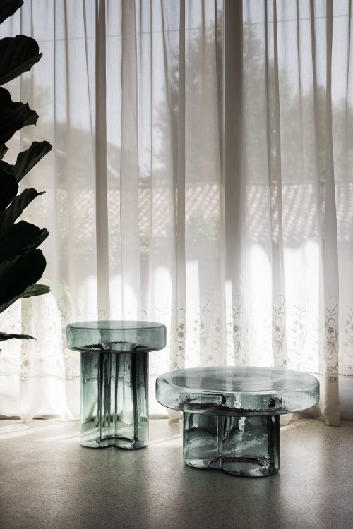 Zwei organisch geformte Beistelltische aus türkisfarbenem Glas stehen vor einem leicht transparenten weissen Vorhang, dahinter ist ein Hausdach zu sehen, links ragt eine Pflanze ins Bild.