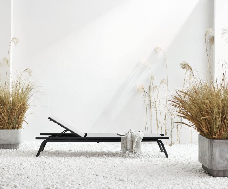 Eine minimalistische schwarze Liege steht vor einem weissen Hintergrund, links und rechts sind zwei Strohpflanzen platziert.