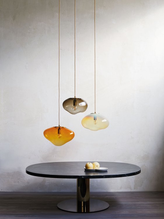 Drei organisch geformte Lampen aus grauem, gelbem und orangem Glas hängen über einem schlichten, schwarzen Tisch mit einem Fussbein.