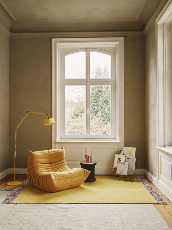 Der gelbe Sessel Togo von Ligne Roset auf einem knallgelben Teppich steht in einem ockerfarben gestrichenen Raum in einer Altbauvilla vor einem Fenster, links über den Sessel beugt sich eine gelbe Leselampe.