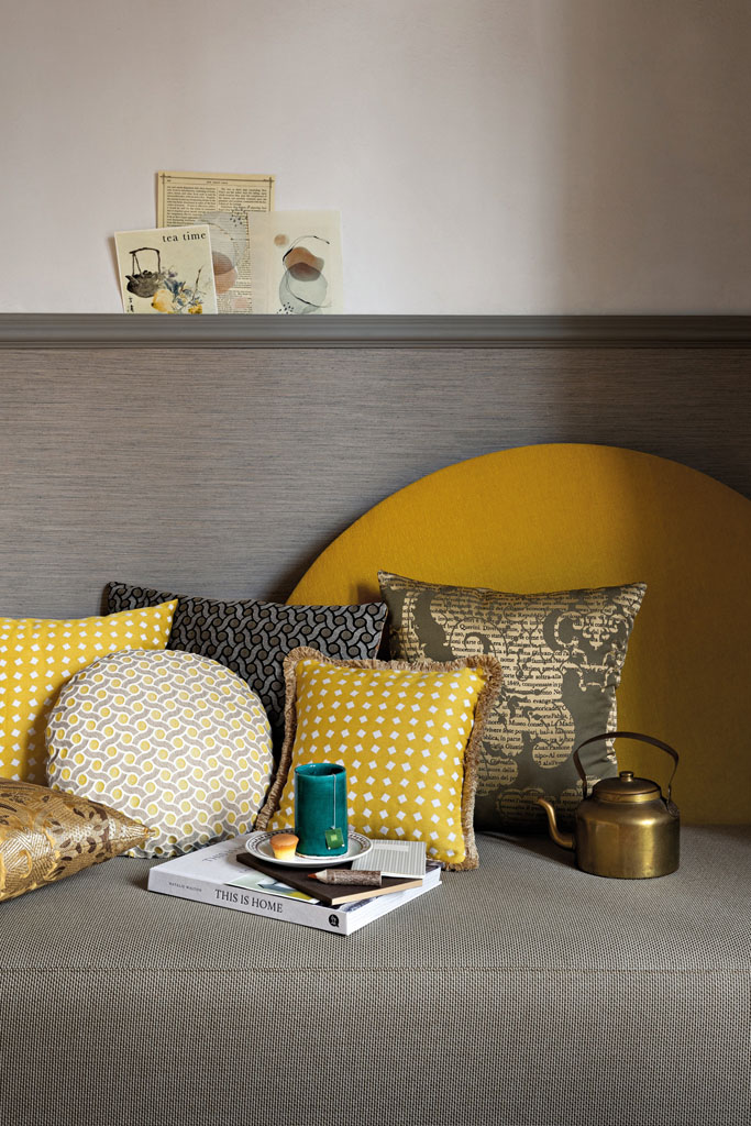 Eine gemütliche Leseecke mit diversen Kissen in Gelb- und Goldtönen auf einem grauen Sofa vor einer grauen Wandvertäfelung.