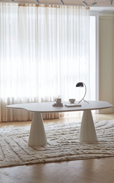 Ein organisch geformter Tisch mit einer runden kleinen Tischlampe steht in einem komplett hell gestalteten Raum vor einem Fenster mit weissen, durchsichtigen Vorhängen.