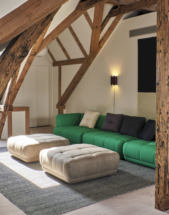 Ein knallgrünes Sofa mit abgerundeten Ecken und abgesteppten Polster steht mit dazu passenden Poufs in hellem Grau-Beige in einem Raum mit Dachstützen und sichtbaren Balken.