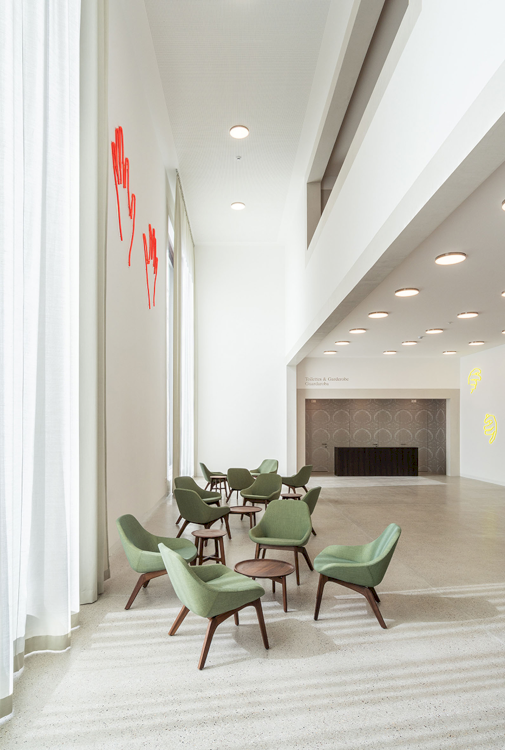 Luftiges und hell gestaltetes Foyer der ner neuen Musikschule Brixen mit weissen Vorhängen, grünen Sesseln, über denen ein rotes Kunstwerk aus Neonröhren hängt.