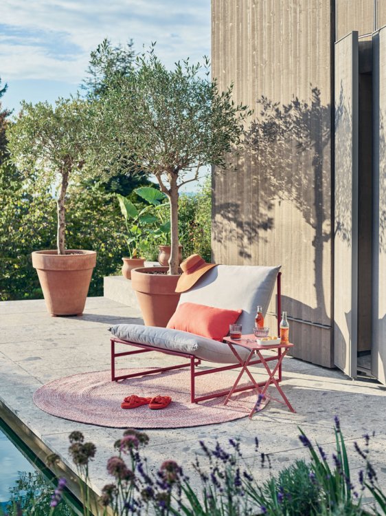 Ein Gartensessel aus feinem roten Metallgestell mit grauem Sitzpolster steht auf einem roten Teppich in einem Garten vor einem Haus mit Holzlattenfassade.
