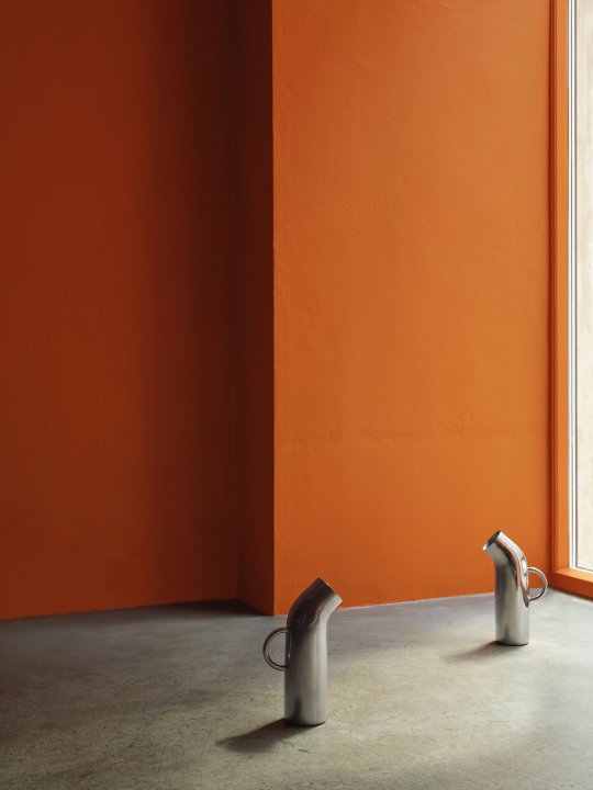 Zwei abgerundete Wasserkaraffen aus Silber in skulpturalem Design stehen am Boden eines Showrooms mit orange gestrichener Wand und vor einem grossen Schaufenster.