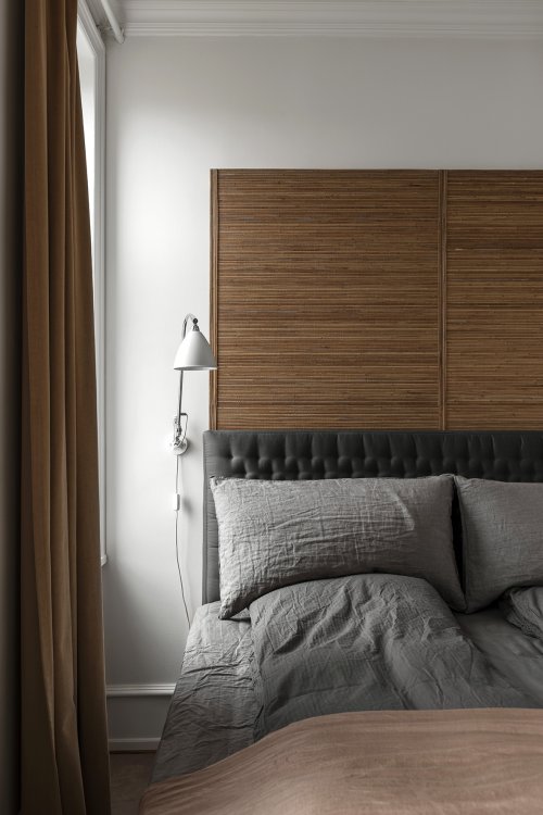 Ausschnitt eines Betts mit grauer Bettwäsche und hohem Kopfteil aus feinen, dunklen Holzlatten das neben einem Fenster mit dunkelbraunem Vorhang steht.