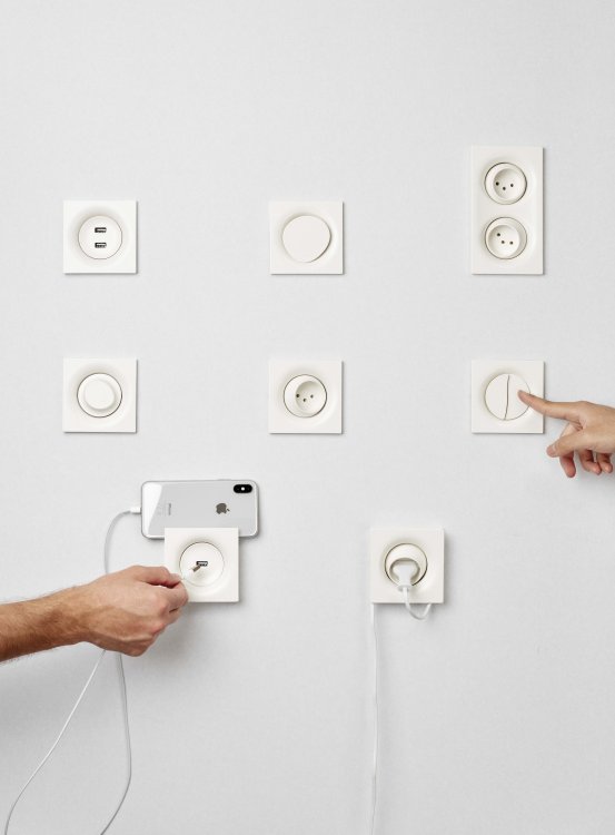 Eine Wand mit verschiedenen Steckdosen und Lichtschaltern mit organisch geformten Tasten an einer Wand, an der zwei Hände gerade dabei sind, ein Kabel einzustecken.