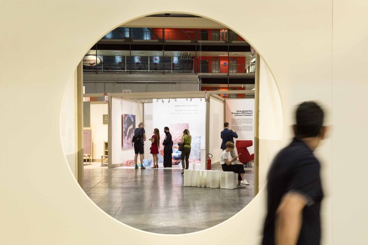 Ein Besucher der Mailänder Möbelmesse Salone del Mobile blickt durch ein rundes Fenster einer hellen Wand, hinter der weitere Besucher des Messegeländes sichtbar sind.