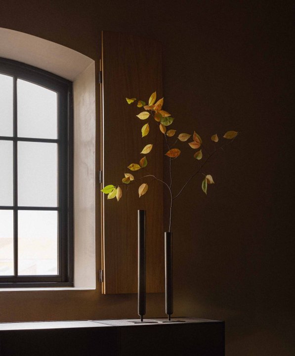 Zwei filigrane Vasen aus dunklem Messing mit Baumzweigen stehen vor einem Fenster.