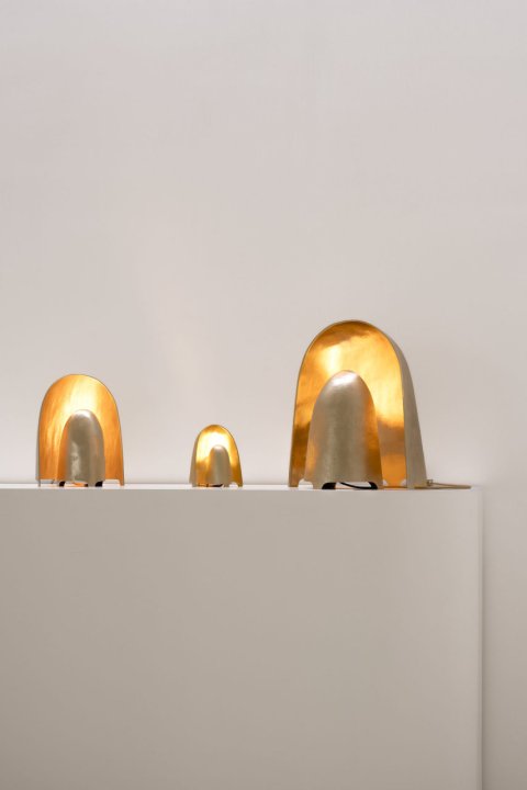Drei unterschiedlich grosse, zweiteilige runde Lampen aus goldener Bronze stehen auf einem beigen Sockel.